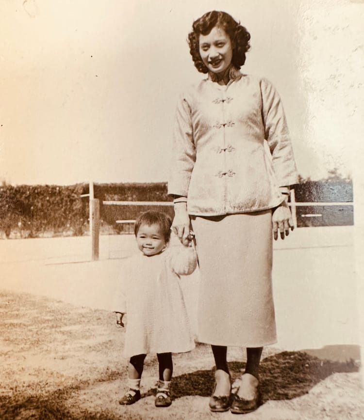 Black-and-white photo of beautiful Chinese woman holding hand of sweet-faced toddler daughter outdoors